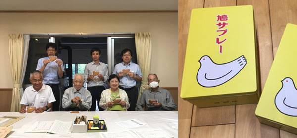 作業に参加した7人が書類を置いたテーブルの後ろに並んで記念撮影。みんなが手には鳩サブレーを持って笑っている。写真の右半分は鎌倉豊島屋の鳩サブレーの黄色い大きな缶が移っている。赤い文字で鳩サブレーと描かれてあって、その下には白い鳩サブレーの形をした鳩が静かに座っているトレードマークが大きく描かれている。