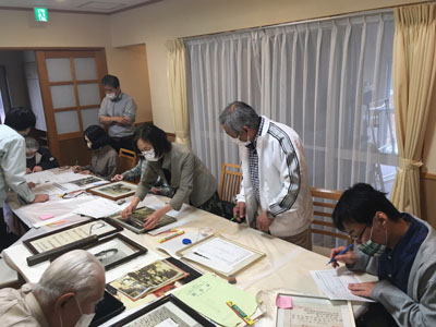 座ったり立ったりしている人々８人が机の周りに居る。史料を１つ１つ見つめながら調査カードを書く者、相談する者の写真