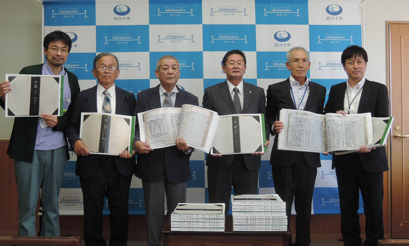 「四万十市」のバックスクリーンの前に整列する関係者一同の記念写真。１人ずつ日誌複製版を掲げている。