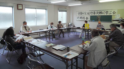 11人が会議室に集まっている。向こうにある黒板には「定期総会」というポスターが貼られている。