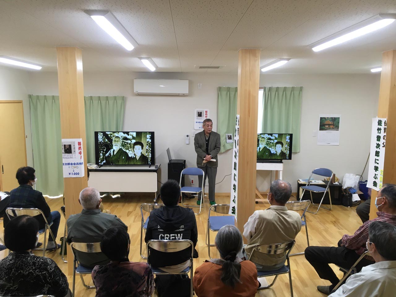 左右２つのモニターの中央に立って話をする中平会長。話しを聞く10名ほどの背中が写っている。