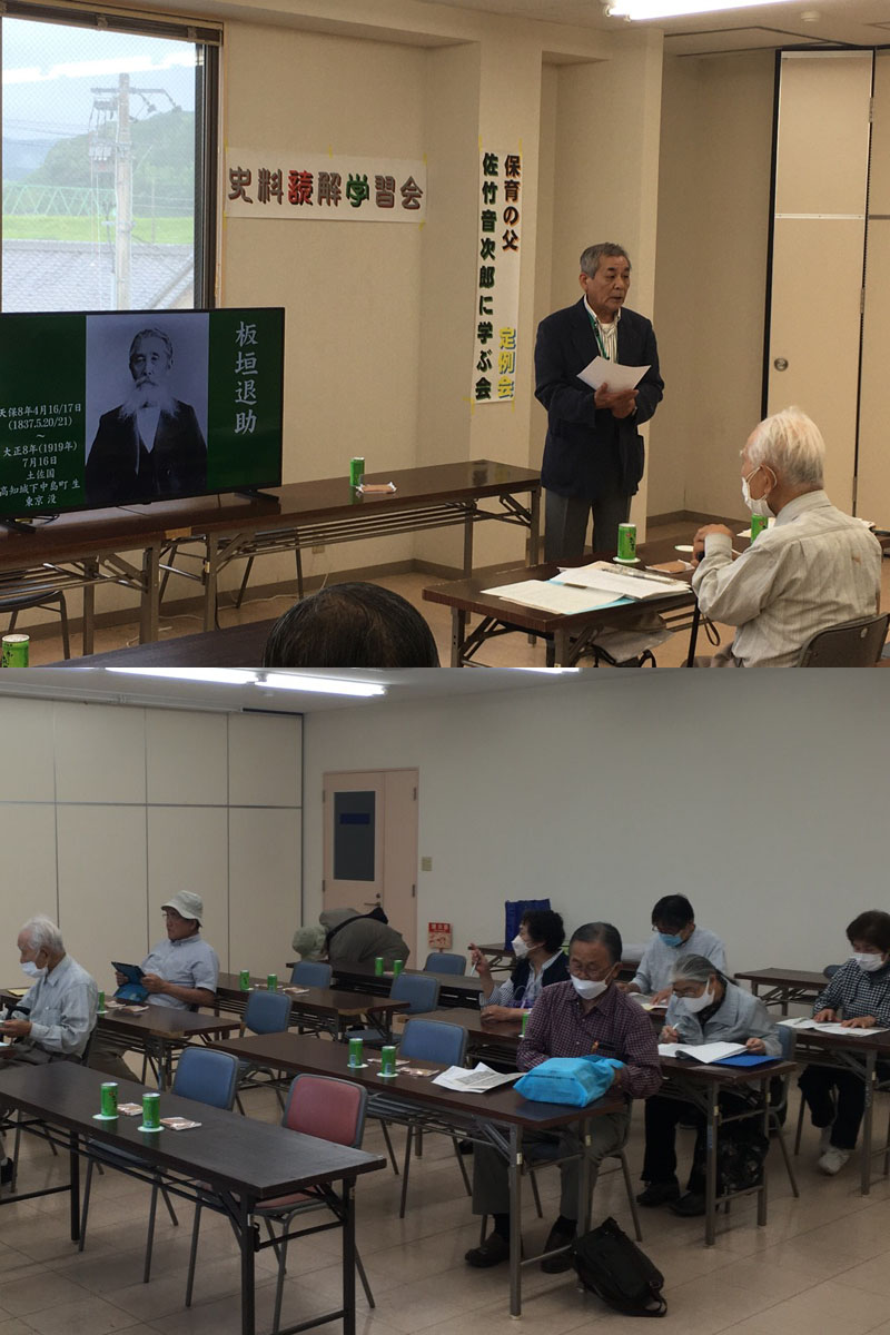 日産サティオ高知四万十店会議室で３回目の読み解き学習会をしている様子。写真上の正面中央には大型液晶モニターが設置されていて板垣退助の肖像が写っている。その右で板垣退助の略歴を説明している中平会長。写真下はスクール形式の会議テーブルに人が座っている。机上には資料と鳩サブレーとお茶。