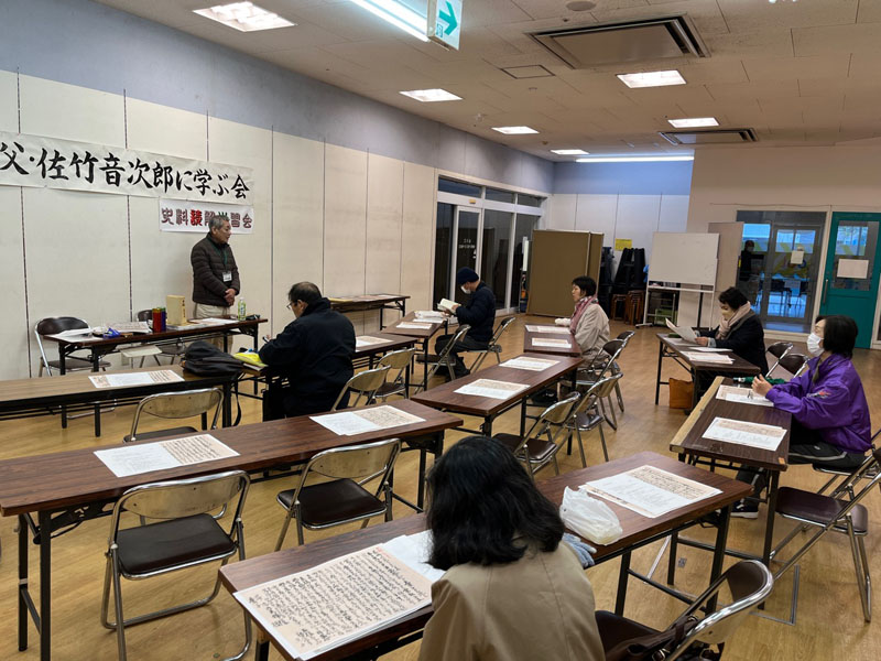 アピアさつきホールで5回目の読み解き学習会をしている様子。弓なりに並んだ会議用テーブルに着席した人々。机上には読み解く原本が並んでいる。中平会長が前に立って説明をしている。