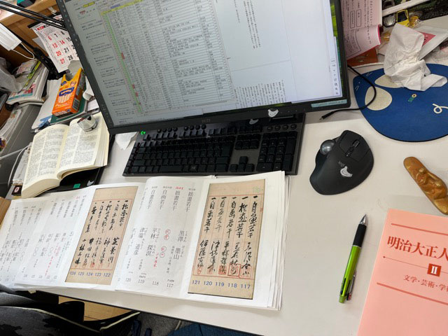 事務用机の上に奉加帳mix版が広げられ、左右に人名事典、ボールペンや文鎮も見える。前にはパソコンとキーボードやトラックボールがあり、画面にはびっしりと文字が並んでいる