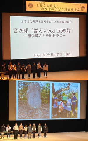 舞台の上で音次郎を発表する竹島小学校５年生。巨大スクリーンには音次郎会が訪問授業した時の様子が映し出されている。