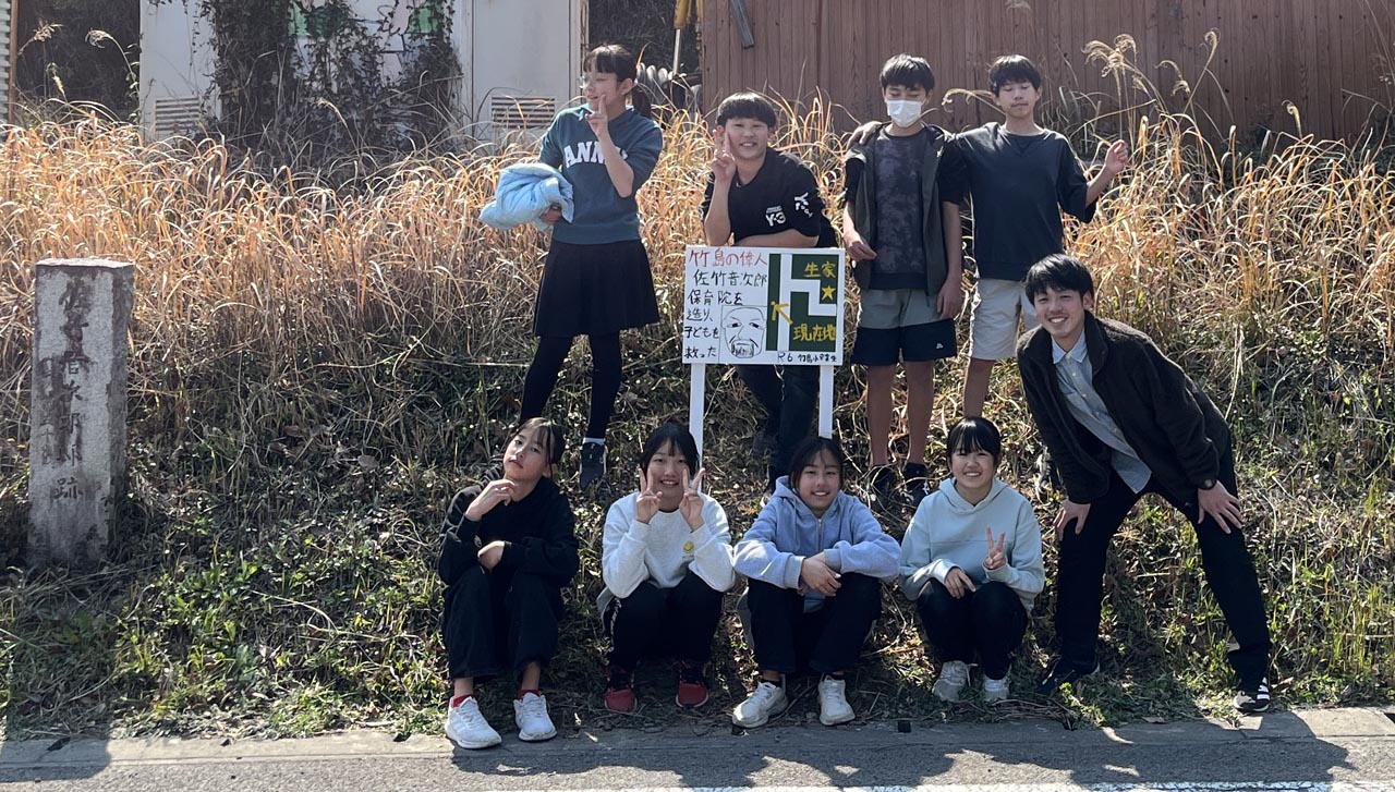 完成した看板と並んで記念撮影。担任の教師と、児童8人が看板を取り囲む。男子3名、女子5名。ピースをしている子供や、斜めを向いてポーズを取るおませさんも居る。